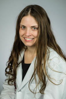 A woman with long brown wavy hair wearing a white lab coat smiles towards the camera